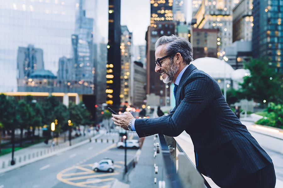 Client Center - Businessman Leans Against a Balcony Wall Overlooking a New York City Street, High Rise Buildings Lighting up in the Background