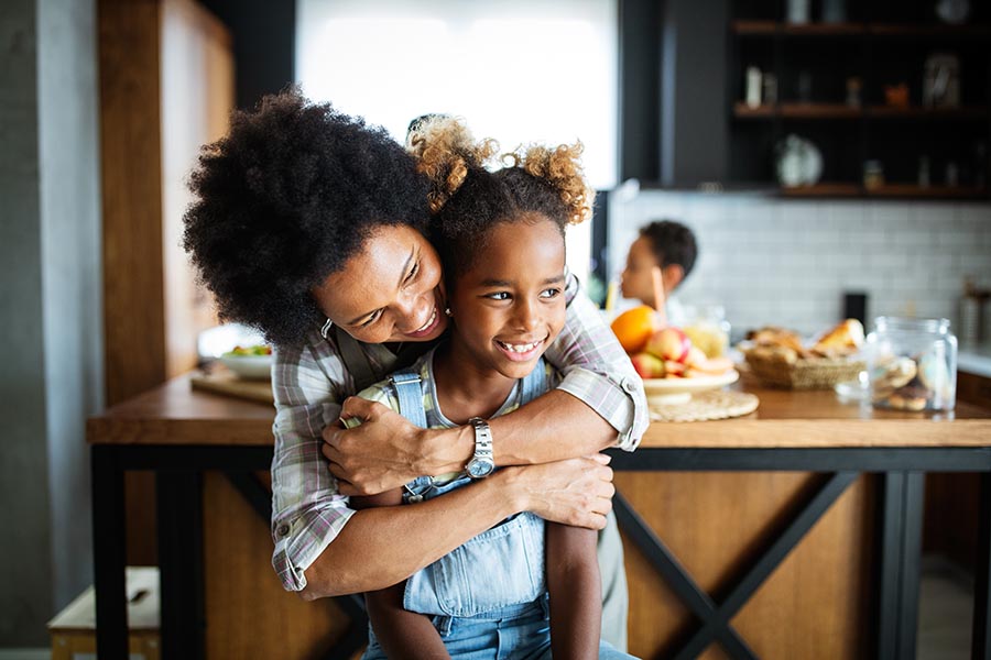 Personal Insurance - Mother Hugs Her Smiling Young Daughter From Behind in Their Kitchen